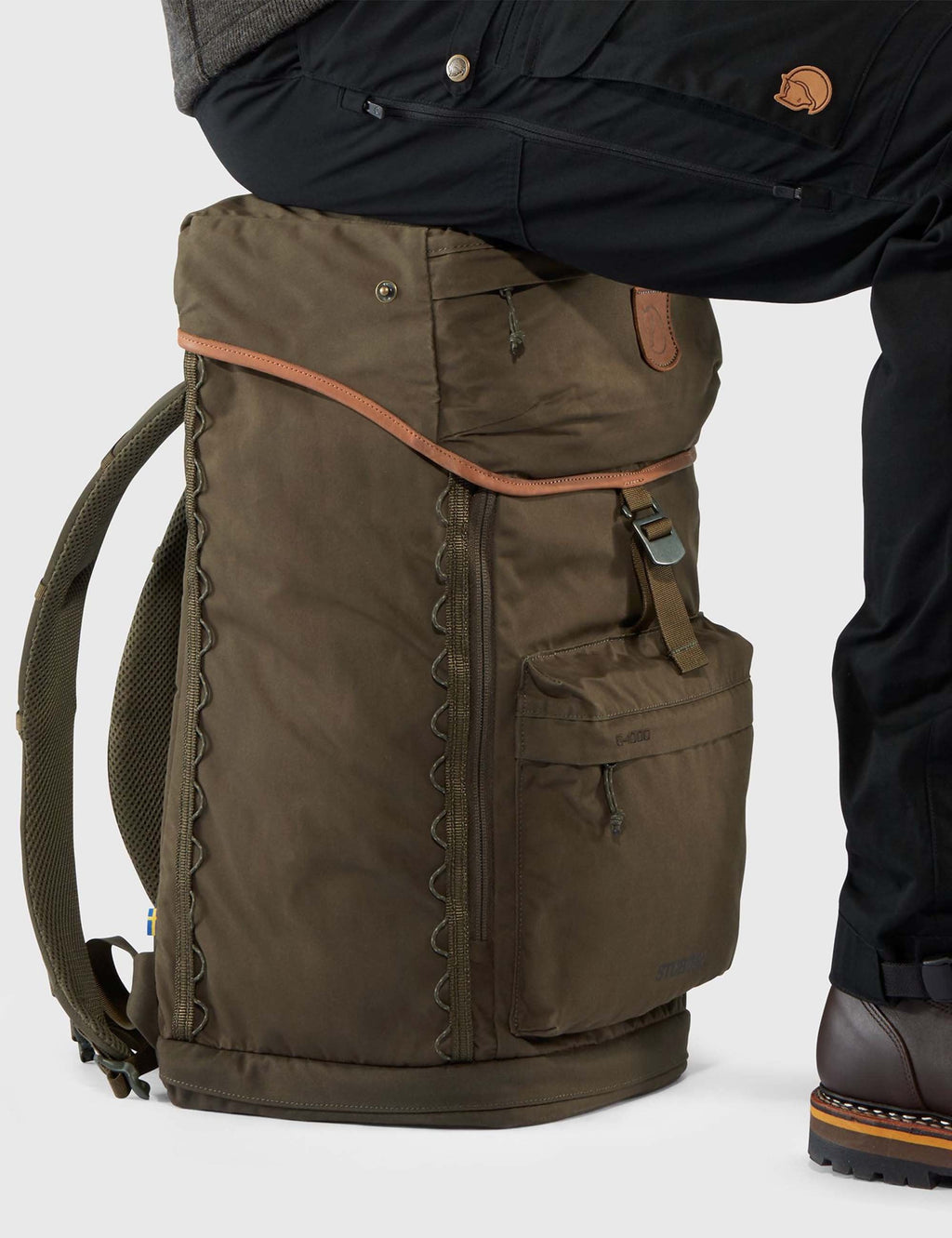Dark olive backpack side view with vertical front panel, zipper, and braided trim.