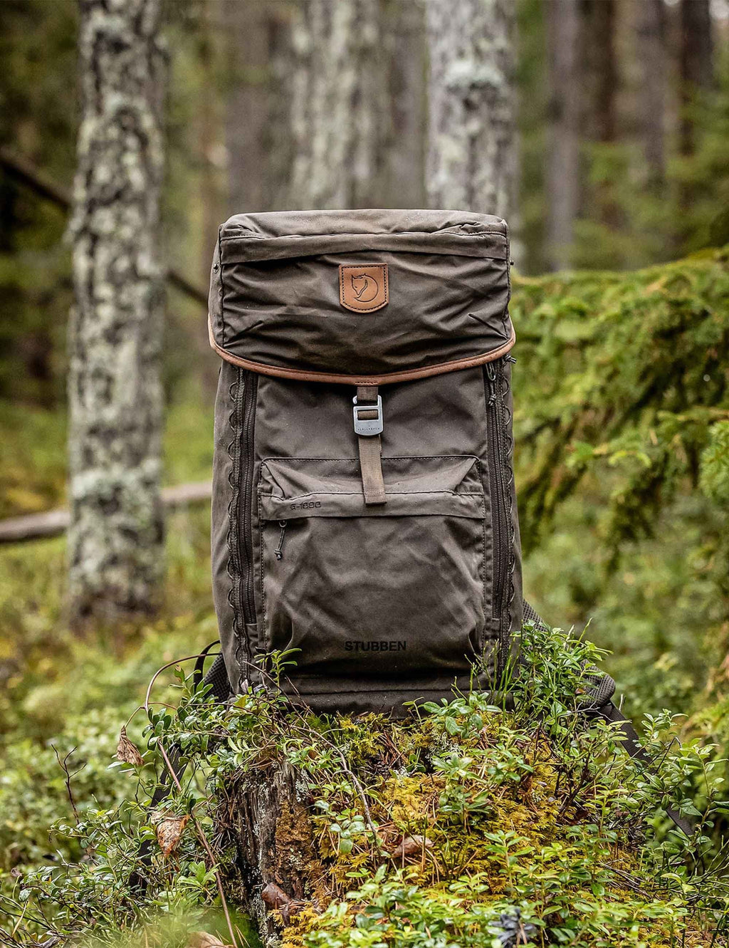 Dark olive backpack with top flap, front logo patch, and two side pockets.