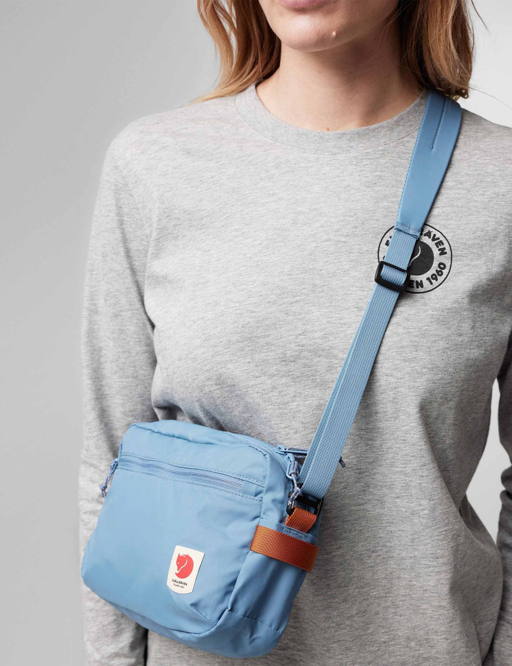 Person wearing a Blue Fjallraven crossbody bag with a visible brand logo on a grey background