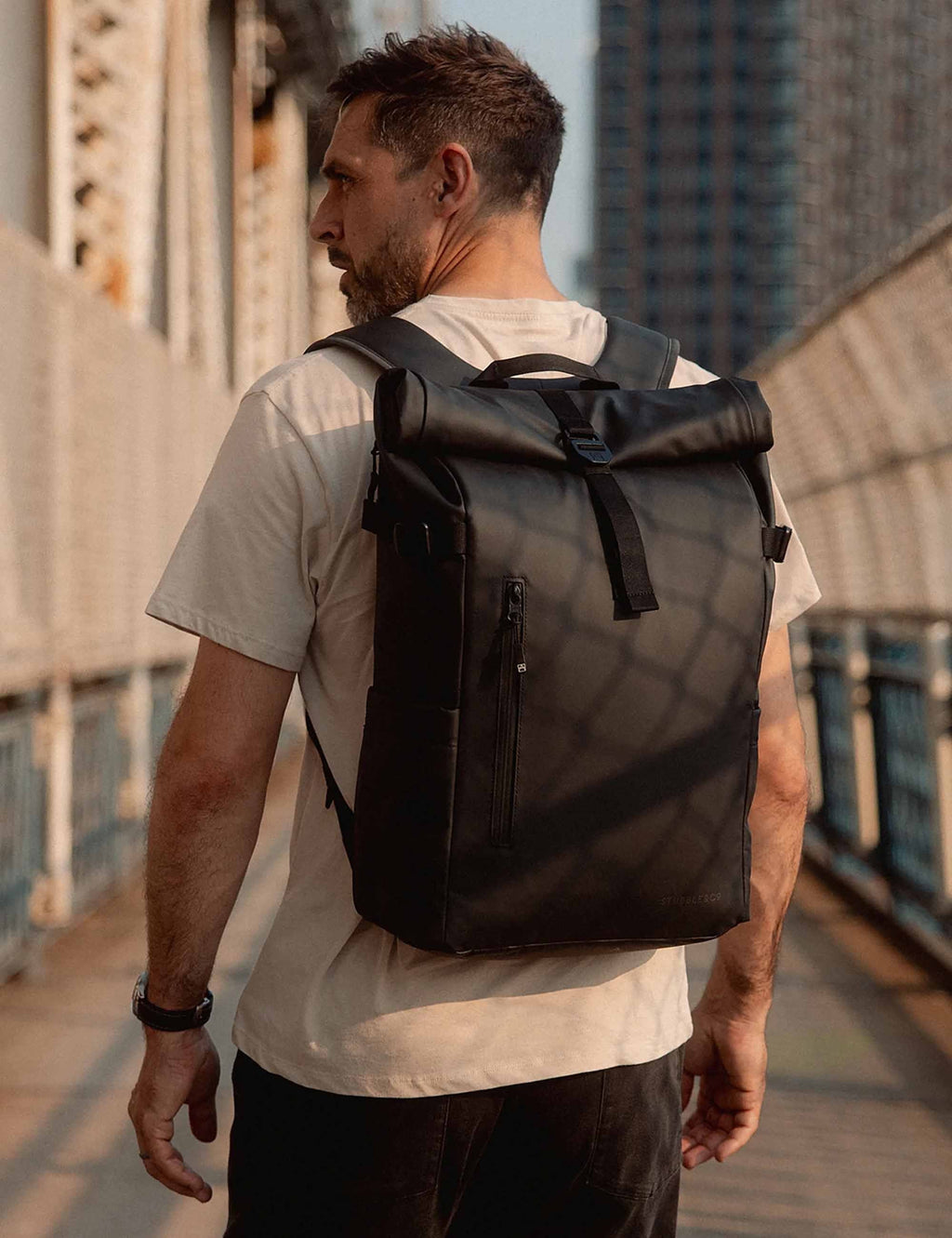All-black roll-top backpack worn by person, with shoulder straps and chest strap