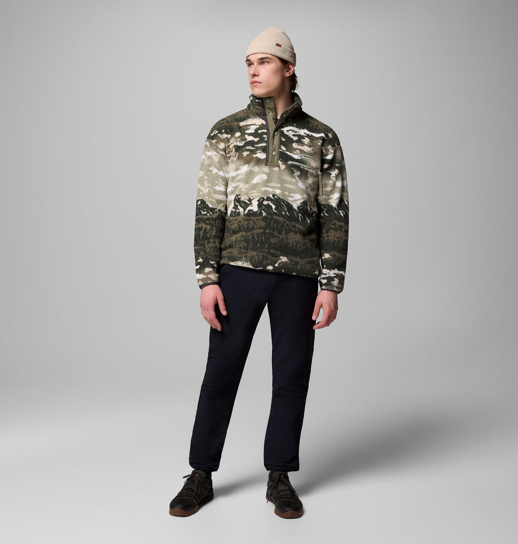 Model wearing khaki mountain-print fleece pullover with stand collar, dark trousers and cream beanie