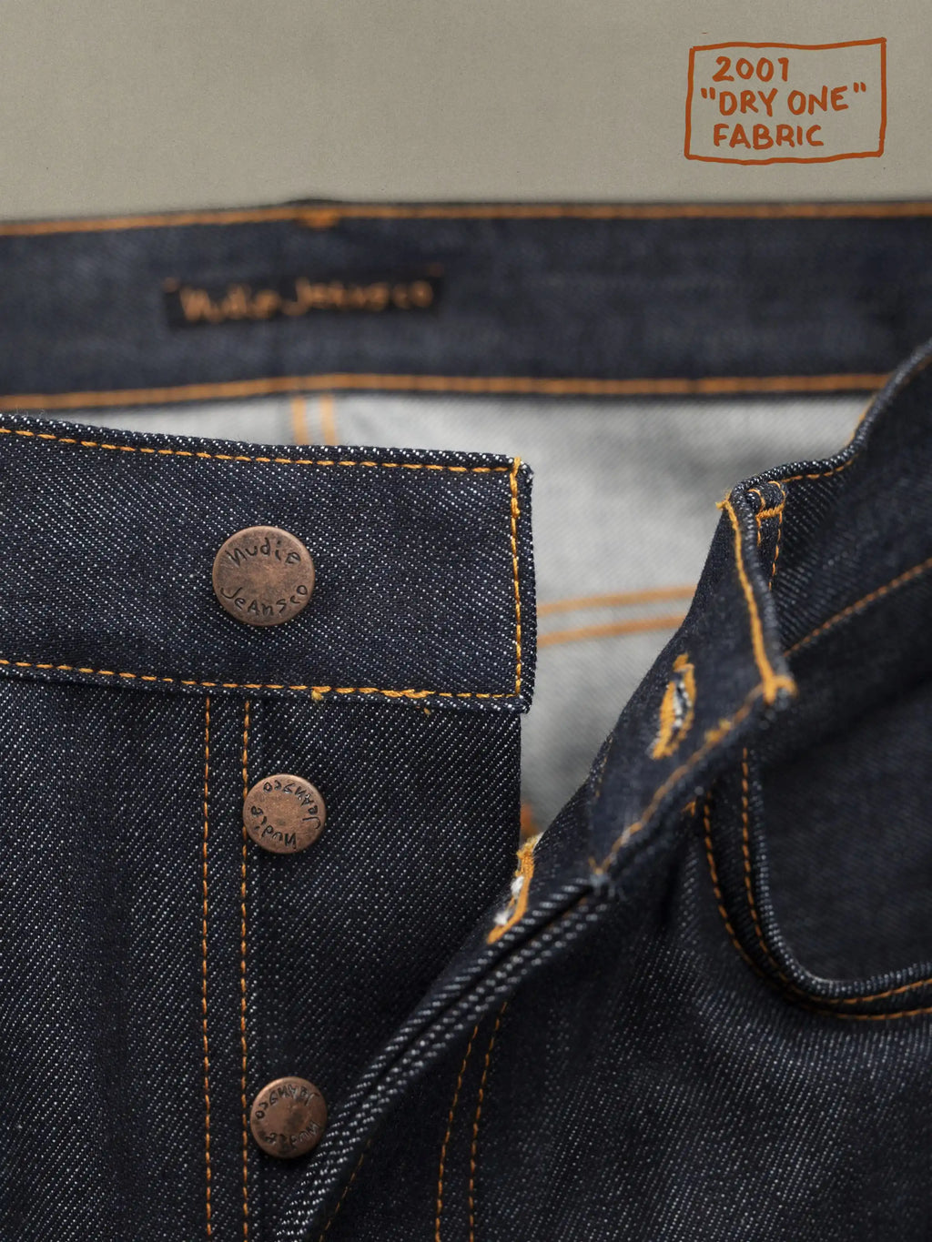 Dark denim waistband and button placket with orange-stitched seams and tan interior label