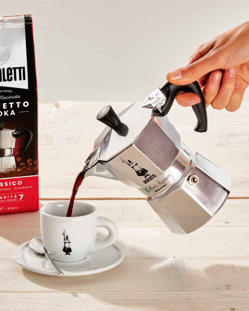 Bialetti Moka pot pouring coffee into a cup with a Bialetti coffee bag in the background.