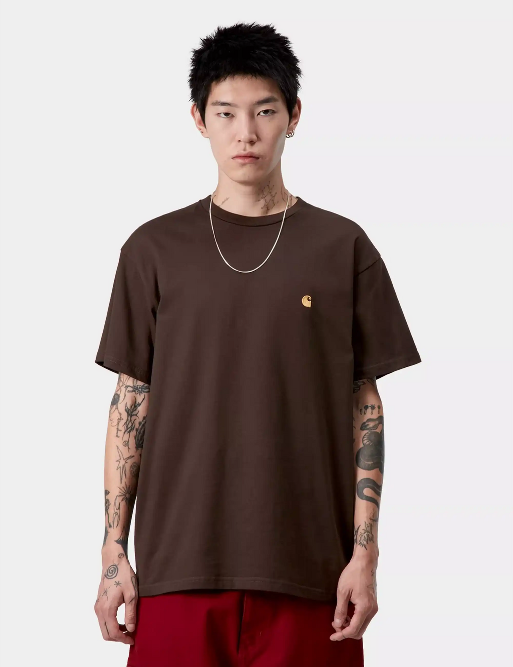 Brown crew-neck T-shirt with small gold chest logo, worn with jeans.