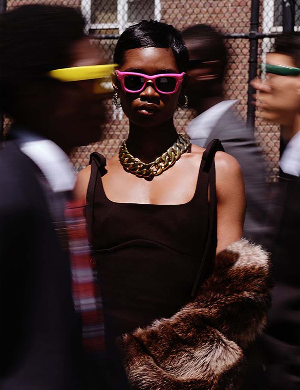 Woman wearing pink sunglasses and a black dress with a fur coat in a crowded area.